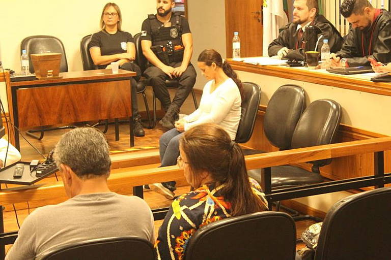 mulher de trança e de branco sentada em tribunal do juri, com policial e promotor e plateia no entorno