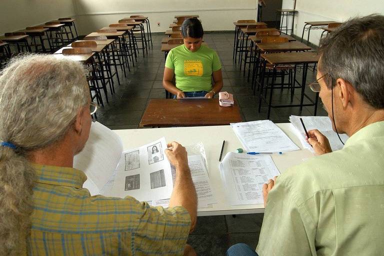Dois professores conferem documentação de estudante, sentada de frente para eles em uma sala de aula