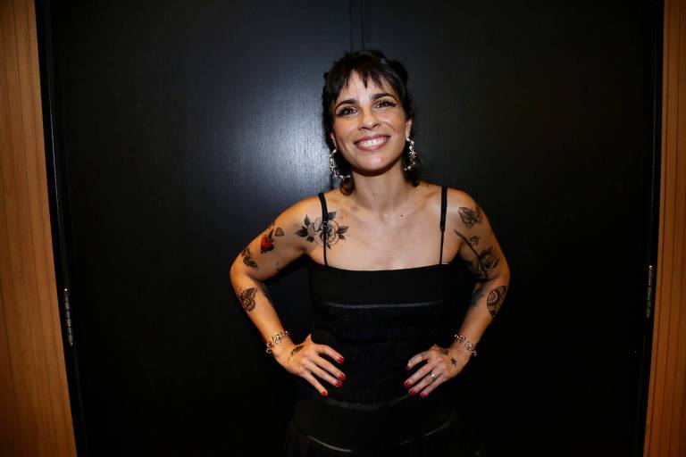     A atriz, escritora e diretora Maria Ribeiro, de vestido preto de alcinhas e parte dos braços cobertos com tatuagens, ri para as câmeras com as mãos na cintura 