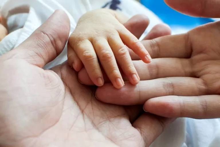 Imagem em close mostra as mãos de um adulto e uma criança recém-nascida