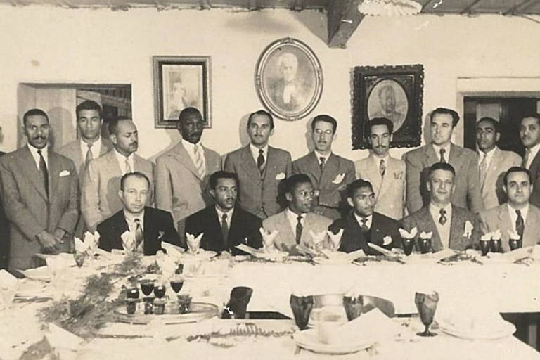 Foto preto e branco antiga traz 18 pessoas de terno. Algumas em pé e outras sentadas à frente. São todos homens, alguns negros e outros brancos.