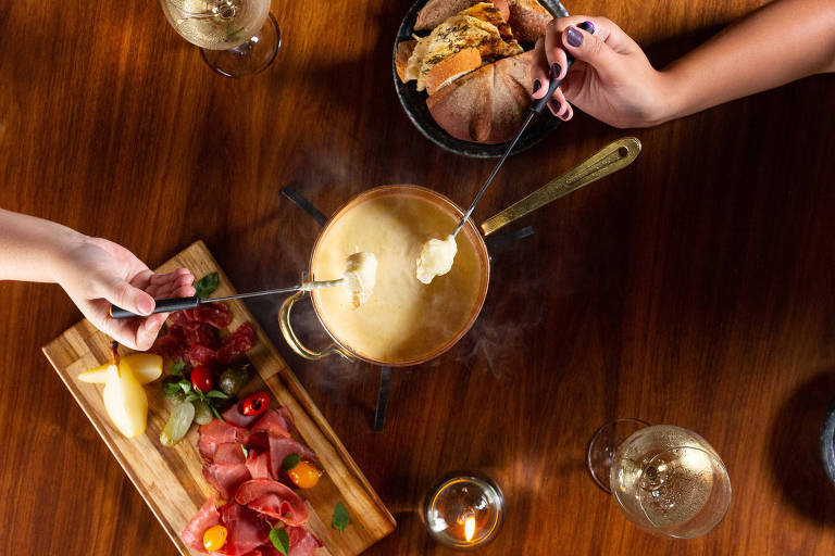 A foto mostra uma mesa com uma panela de fondue de queijo e duas mão segurando espetos mergulhados no fondue