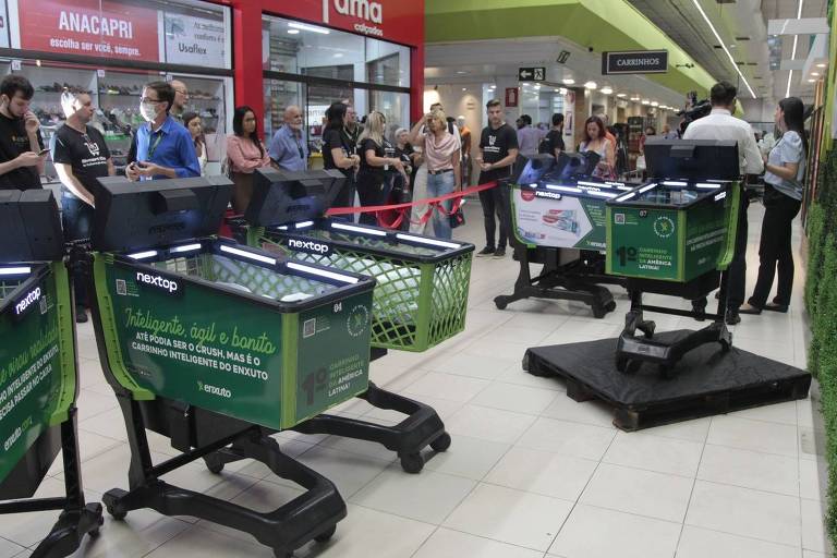 Imagem mostra quatro carrinhos de supermercado, nas cores preto e verde, dentro de um centro de compras. Ao redor, pessoas olham para eles.