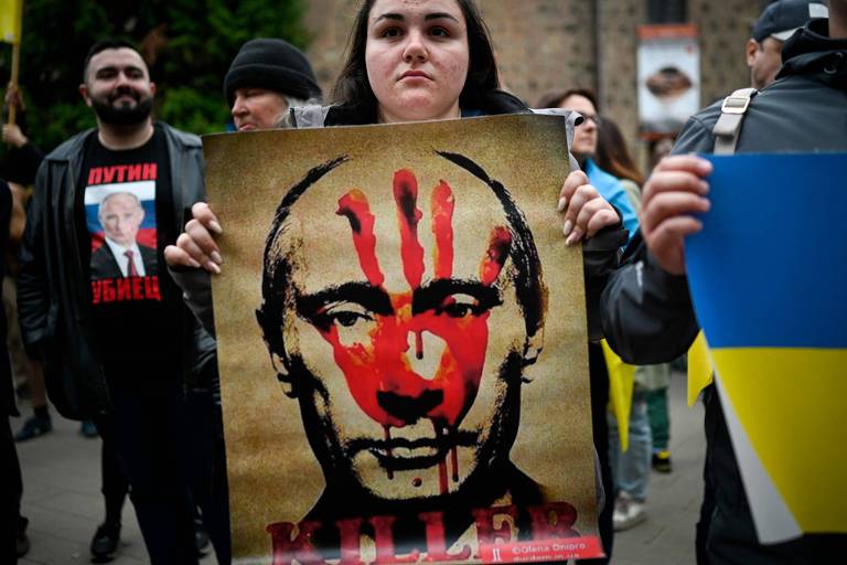 Mulher segura cartaz que traz o desenho de uma mão vermelha cobrindo o rosto de Vladimir Putin, tendo ao lado uma bandeira da Ucrânia em amarelo e azul.