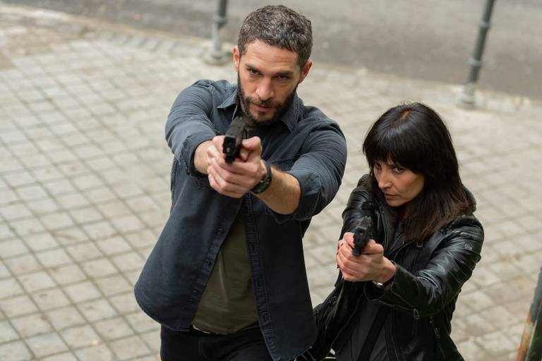 Cena de série retratando um homem e uma mulher brancos vestidos casacos pretos e empunhando pistolas em direção a um alvo não mostrado na imagem