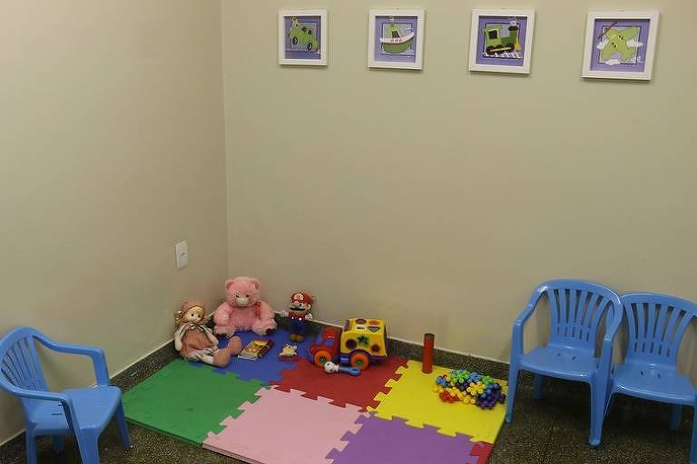sala com tapete colorido infantil, urso e brinquedos, com três cadeiras de plástico azuis
