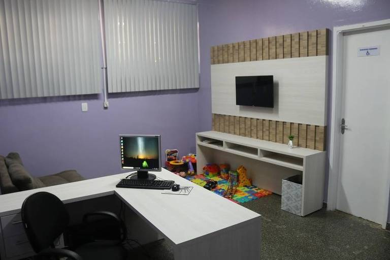 sala com uma mesa com computador, cadeira, um armario ao fundo e um tapet infantil com brinquedos