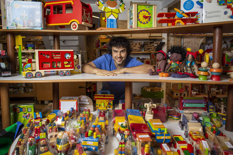 Homem branco de camiseta azul em meio a brinquedos de madeira coloridos e bonecas de feltro negras