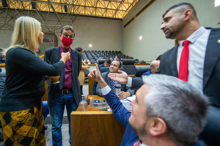 Uma mulher loira e quatro homens de terno e gravata debatem apontando os dedos uns para os outros em plenário.