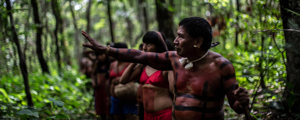 Indígenas caminham em fila em uma área de floresta. O cacique, o primeiro da fila, está com o braço direito erguido, apontando para algum lugar da floresta