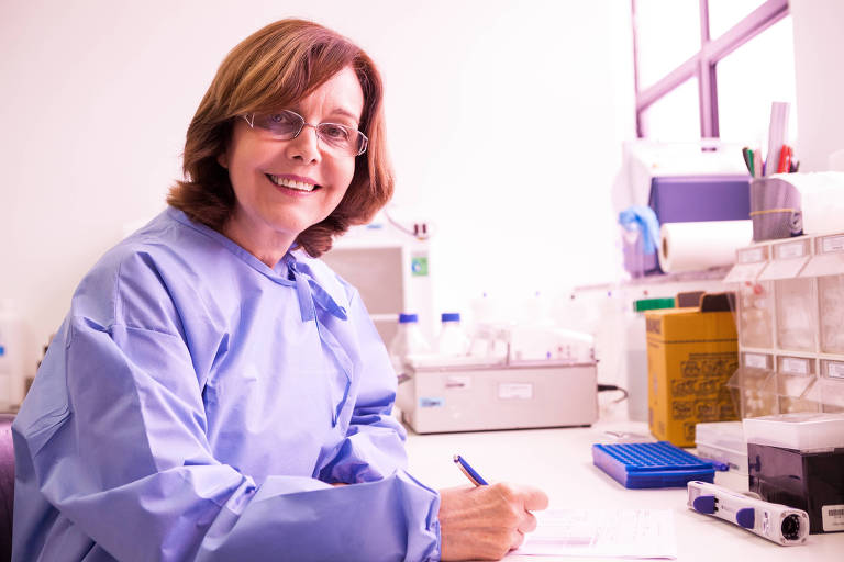 Marilda Siqueira usa um avental e faz anotações em um papel, na mesa de um laboratório