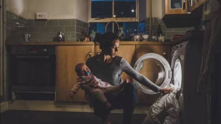 Mãe segura filho no colo enquanto coloca roupas dentro da máquina de lavar