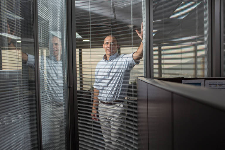 Imagem mostra Pedro Zinner, um homem branco que veste camisa azul e calça branca, se apoiando numa janela de vidro, dentro de um escritório.