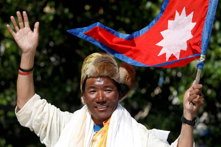 Homem de trajes brancos e chapeu de pelo animal segura bandeira do Nepal, vermelha com desenhos brancos