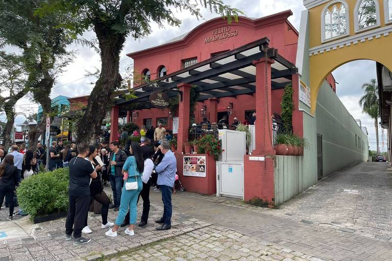 Imagem mostra multidão de pessoas reunida em frente a um restaurante vermelho. Há pessoas na calçada e na varanda do estabelecimento. Do lado direito, há uma rua de paralelepípedo.