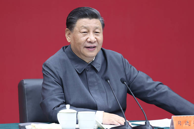 Em fundo vermelho, vê-se o dirigente chinês sentado em frente a uma mesa em que estão um microfone e xícaras de café