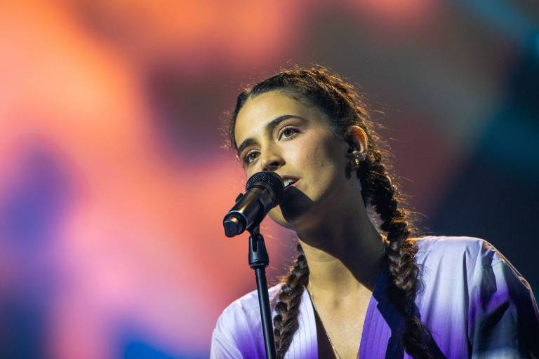 Imagem em close-up da jovem cantora Maro, branca e de cabelos longos penteados em duas tranças, em frente a um microfone durante ensaio para apresentação. no festival Eurovision, onde representa Portugal