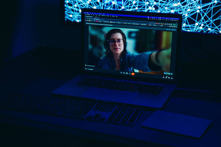Uma foto azul escura mostra uma videochamada em um notebook; uma mulher branca de cabelo longo castanho está na tela