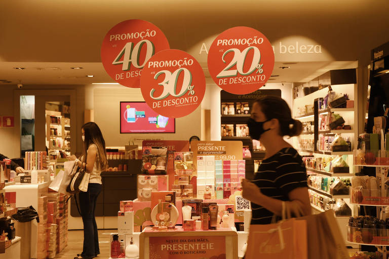 Imagem mostra mulher com sacolas de compra passando em frente a uma loja de shopping.