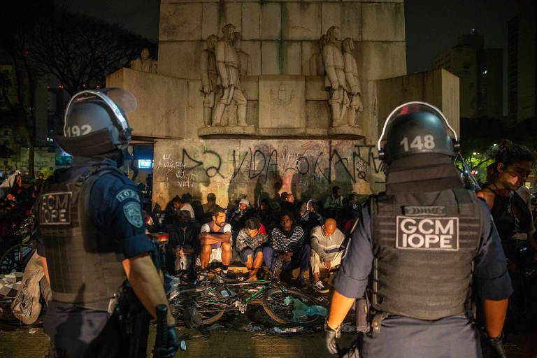 Dois policiais da GCM vigiam um grupo grande de pessoas sentadas ao pé de um monumento pixado, de noite.