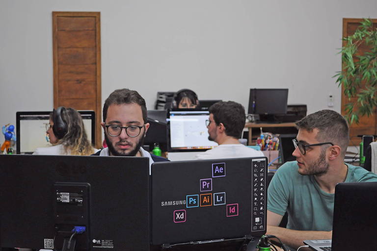 dois homens estão em frente a computadores, com mulheres atrás de costas, em um escritório