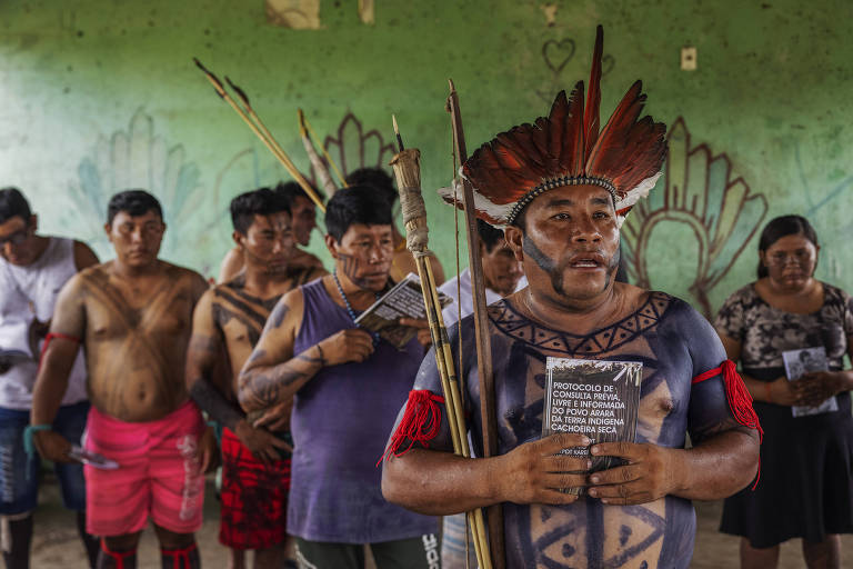 Homem com cocar e pinturas corporais segura com as duas mãos um livreto; atrás dele estão outros indígenas