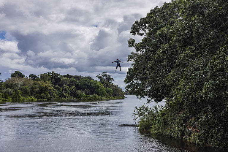 Com os braços e pernas abertos corpo de menino flutua no ar perto da copa de uma árvore, em direção ao rio
