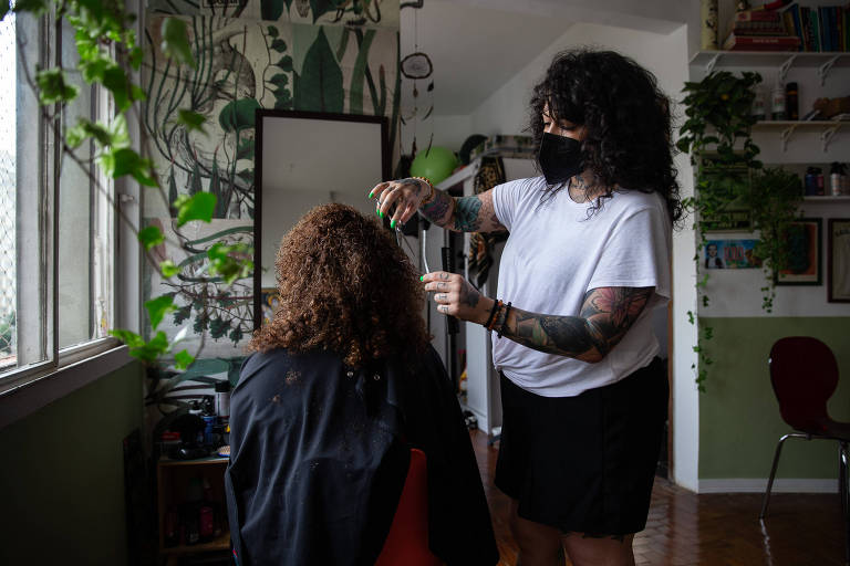Imagem mostra mulher branca de cabelo cacheado, com camisa branca e saia preta, cortando cabelo ruivo e cacheado de uma mulher que está de costas para a câmera. O ambiente é a sala de uma casa, com paredes brancas e verdes, plantas e espelho.