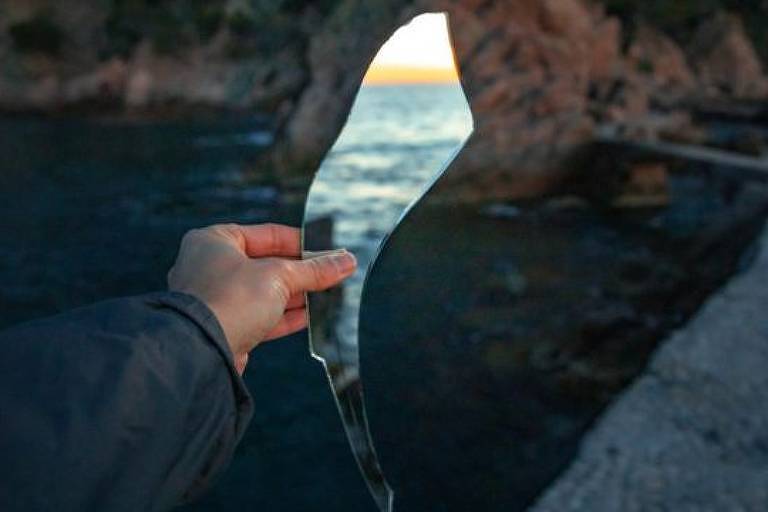 Imagem em close mostra a mão de uma pessoa segurando um pedaço quebrado de espelho, que reflete a imagem de um mar