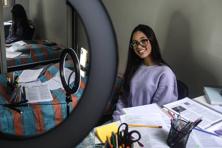 Na foto, vemos Jessica Ellen Silva, 20, sentada em frente a uma luz de led circular e com o celular em um tripé. AL lado, está uma mesa com materiais de estudo em cima, folhas, lápis e cadernos

