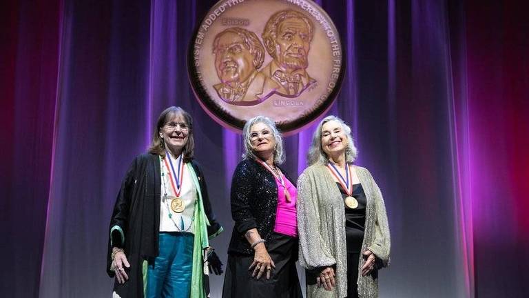 três mulheres brancas e idosas em palco iluminado por cores arroxeadas