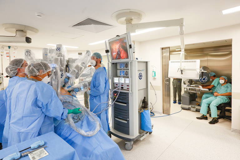 Imagem colorida mostra vários profissionais médicos  em uma sala de cirurgia. Eles vestem roupa azul, com touca azul, e estão em volta de uma mesa cirúrgica. No lado direito, ao fundo, médico vestindo roupa verde  manuseia uma máquina.