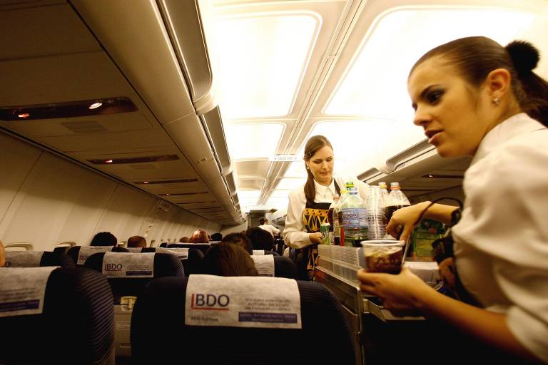 Duas mulheres levam carrinho e servem bebidas e comidas a passageiros em corredor de avião. 