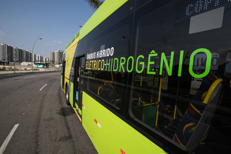 Imagem mostra ônibus verde numa avenida ao lado de um condomínio de prédios. Na janela do ônibus está escrito "Ônibus híbrido: elétrico-hidrogênio"