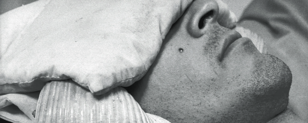 Retrato em preto e branco de homem branco com almofada cobrindo seus olhos e conchas tapando os ouvidos
