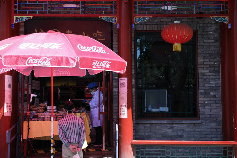 Homem de costas é visto esperando encomenda do lado de fora de restaurante; ao fundo funcionário trabalha e ao lado, fachada da loja com lanterna vermelha, tipicamente chinesa 