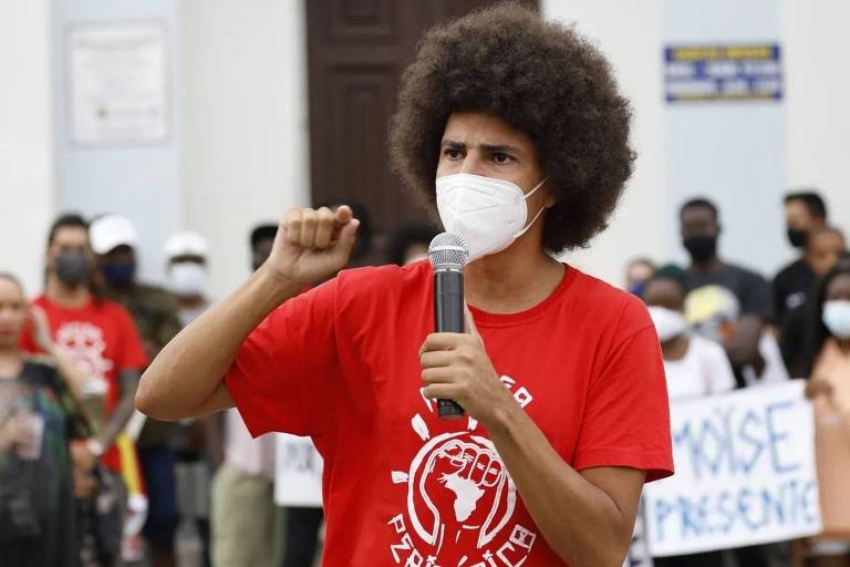 homem de máscara e punho erguido fala ao microfone em igreja cercado de membros de protesto