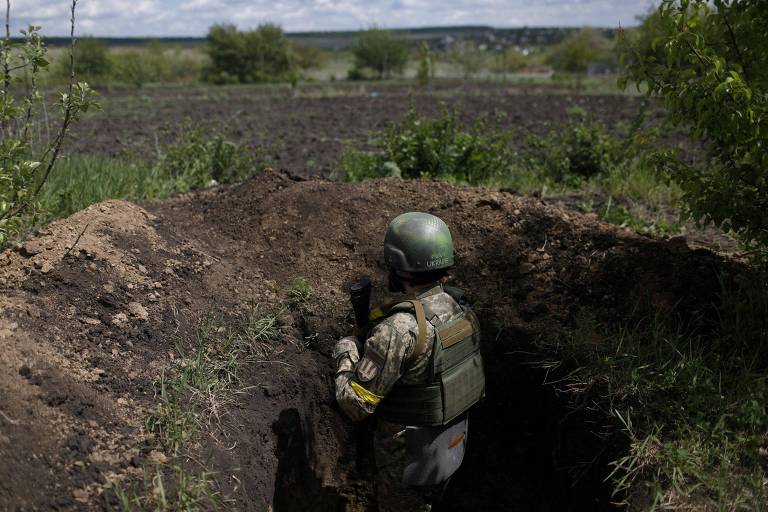 Soldado é visto de costas dentro de uma trincheira; buraco de terra encobre o oficial em meio a um campo aberto com vegetação 