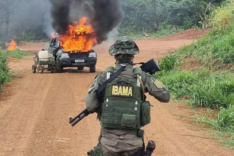 Imagem em primeiro plano mostra uma pessoa de costas com o uniforme do Ibama e um fuzil na mão. Ela está no meio de uma estrada de terra. Ao fundo, se vê um carro em chamas.
