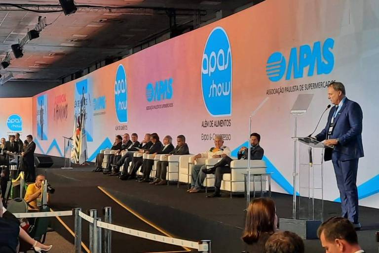 João Galassi, presidente da Abras, aparece em primeiro plano em um palco. Ele está em pé, usando terno na cor azul. Ao fundo, estão sentadas outras autoridades, incluindo o presidente Jair Bolsonaro. Ele discursa para uma plateia, em sua frente.