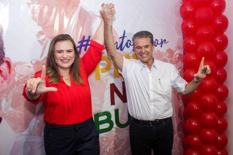 Marília Arraes sorridente vestindo camisa vermelha, com a mão direita fazendo a letra L alusiva ao ex-presidente Lula e o outro braço erguido junto com o braço de André de Paula, que veste camisa branca e sorri fazendo também o L em alusão a Lula com os dedos da mão esquerda.