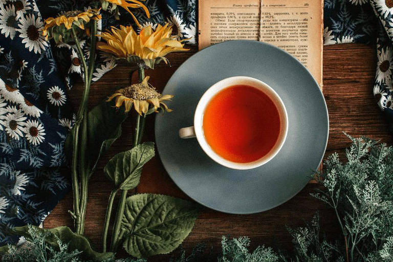 Imagem de um chá de cor avermelhada em uma mesa decorada com flores e com um livro