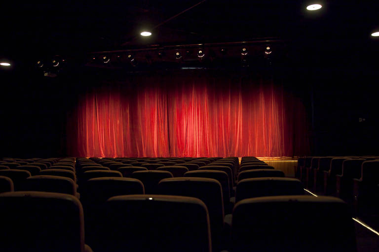 Vista de trás do espaço interno do teatro com as poltronas vermelhas, palco ao fundo e uma grande cortina vermelha