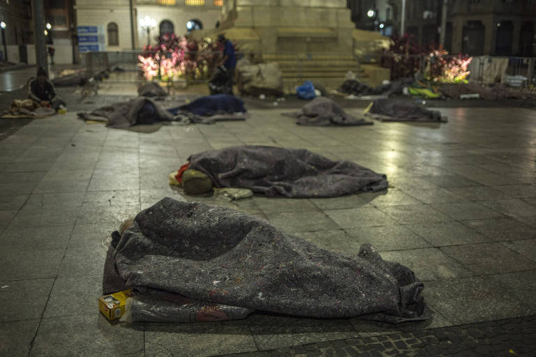 Pessoas deitadas no chão, enroladas em cobertores ao pé de monumento