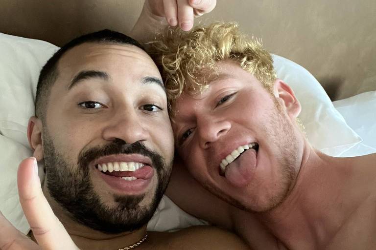 Dois homens sem camisa posando para selfie deitados em uma cama