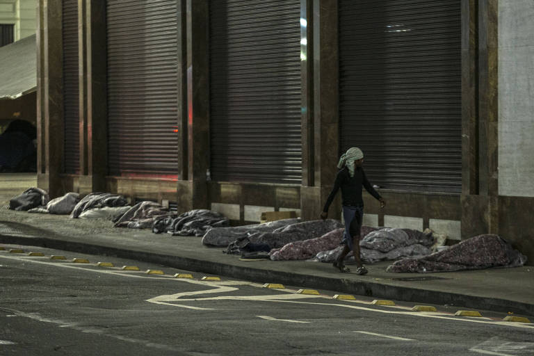 Pessoas dormem em calçada enroladas em cobertores cinza enquanto um homem caminha pela rua