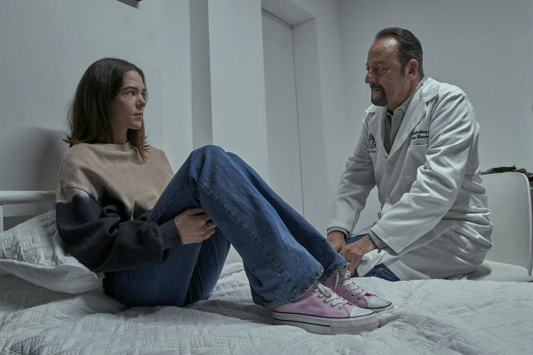 Cena de série em que uma menina de blusa bege e calças jeans está sentada na cama de um hospital diante de um homem calvo de jaleco branco