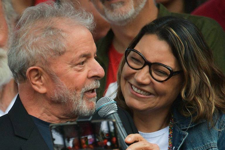 Imagem mostra Janja, uma mulher branca, de cabelo castanho e loiro, segurando o microfone para Lula, enquanto ele discursa. Ela está usando uma jaqueta jeans, camiseta branca e óculos pretos. Eles estão cercados de apoiadores.
