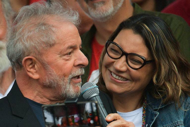 Lula fala ao microfone e Janja, ao lado, sorri