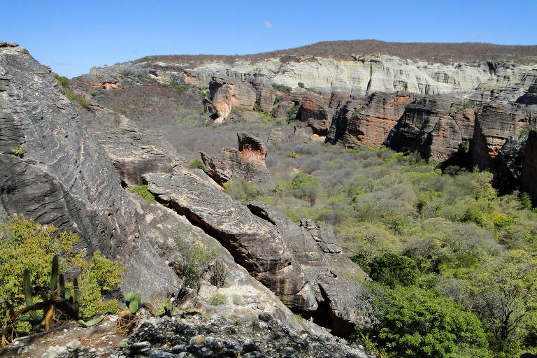 Imagem colorida, em registro feito durante o dia, mostra um morro ao fundo, e o céu azul. À frente algumas rocas, cercadas de vegetação.